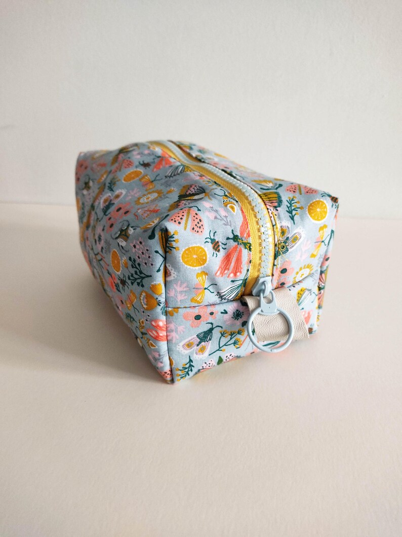 May include: A light blue zippered pouch with a floral print featuring butterflies, flowers, and lemons. The zipper has a yellow pull tab and a light blue ring.