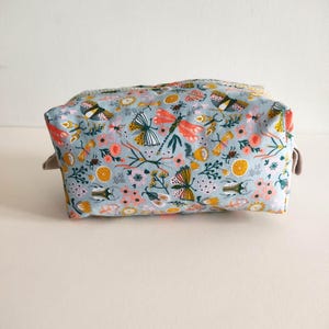May include: A light blue toiletry bag with a floral pattern featuring butterflies, dragonflies, and other insects. The bag has a zipper closure and a beige fabric lining.