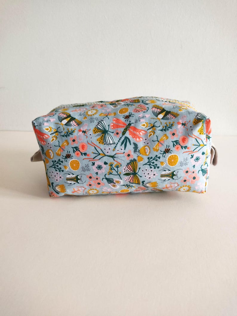 May include: A light blue toiletry bag with a floral pattern featuring butterflies, dragonflies, and other insects. The bag has a zipper closure and a beige fabric lining.