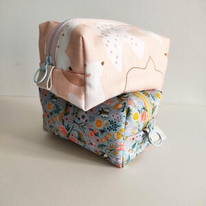 May include: Two fabric zippered pouches stacked on top of each other. The top pouch is pink with a white abstract pattern. The bottom pouch is light blue with a floral pattern.