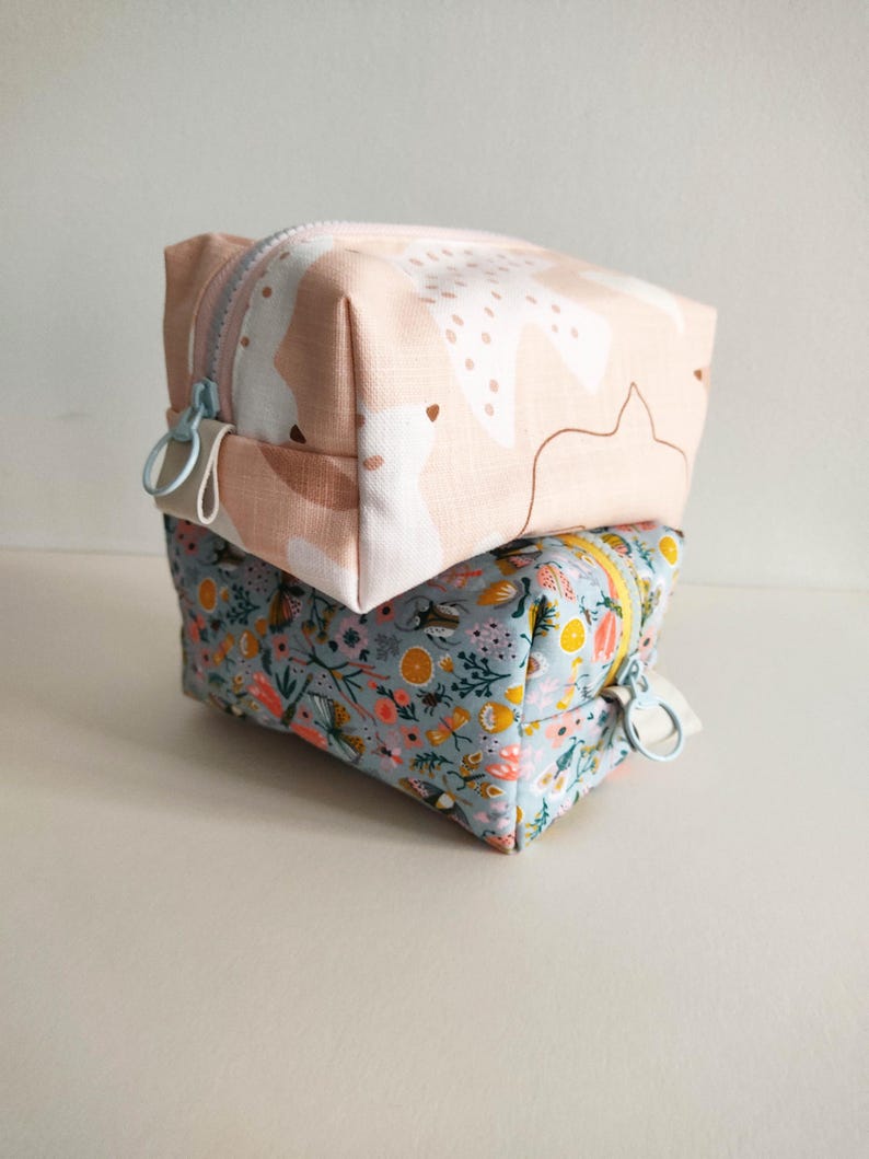 May include: Two fabric zippered pouches stacked on top of each other. The top pouch is pink with a white abstract pattern. The bottom pouch is light blue with a floral pattern.