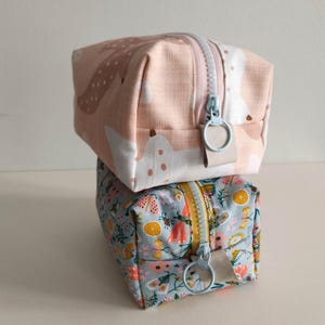 May include: Two fabric zippered pouches stacked on top of each other. The top pouch is pink with a white and brown abstract pattern. The bottom pouch is blue with a floral pattern. Both pouches have light blue zipper pulls.