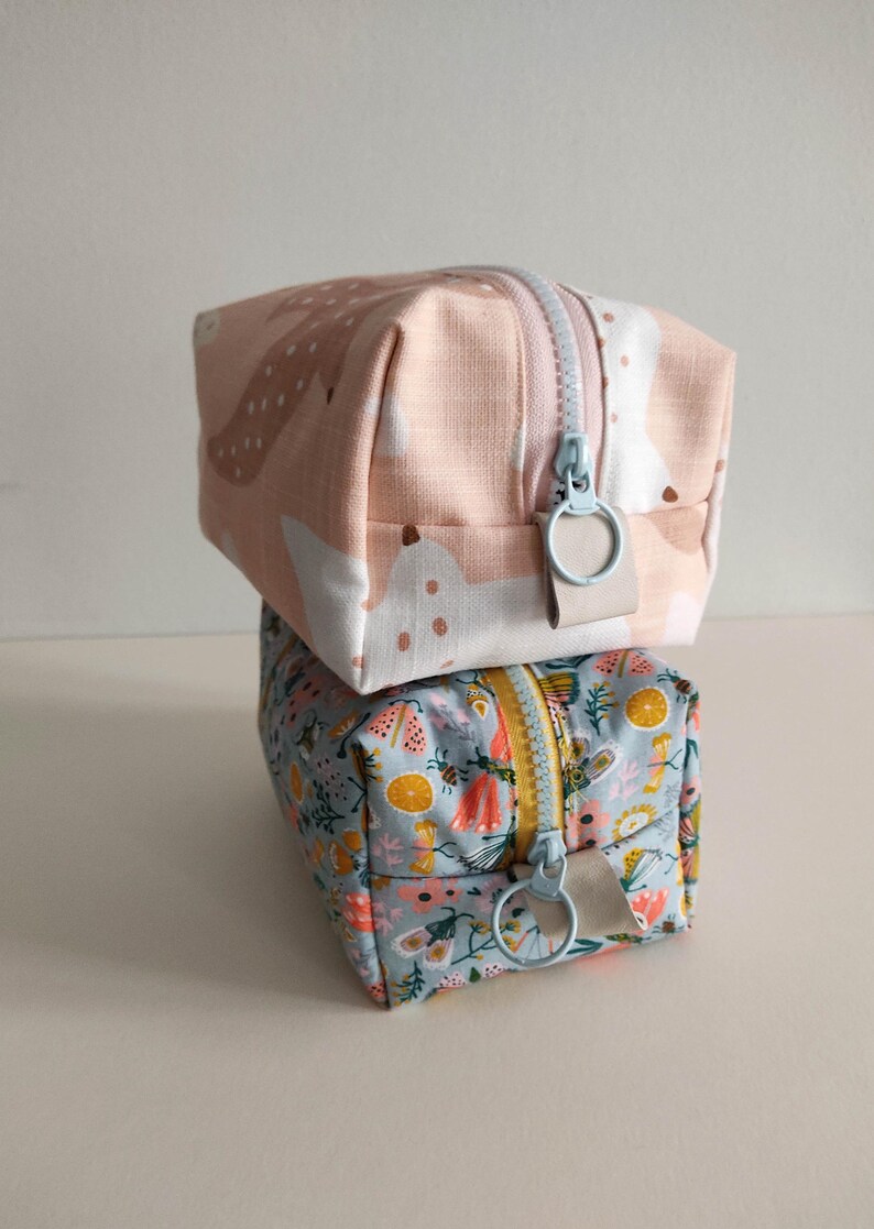 May include: Two fabric zippered pouches stacked on top of each other. The top pouch is pink with a white and brown abstract pattern. The bottom pouch is blue with a floral pattern. Both pouches have light blue zipper pulls.