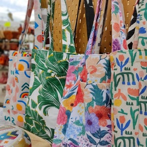 Może przedstawiać: Pięć toreb typu tote z różnymi wzorami kwiatowymi i geometrycznymi. Torby wiszą na tablicy z otworami o jasnobrązowym tle. Torby są wykonane z jasnego materiału i mają różne kolory, w tym zielony, różowy, niebieski i żółty.