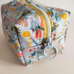May include: A light blue zippered pouch with a floral and insect print. The pouch has a yellow zipper and a light blue ring attached to a beige strap.