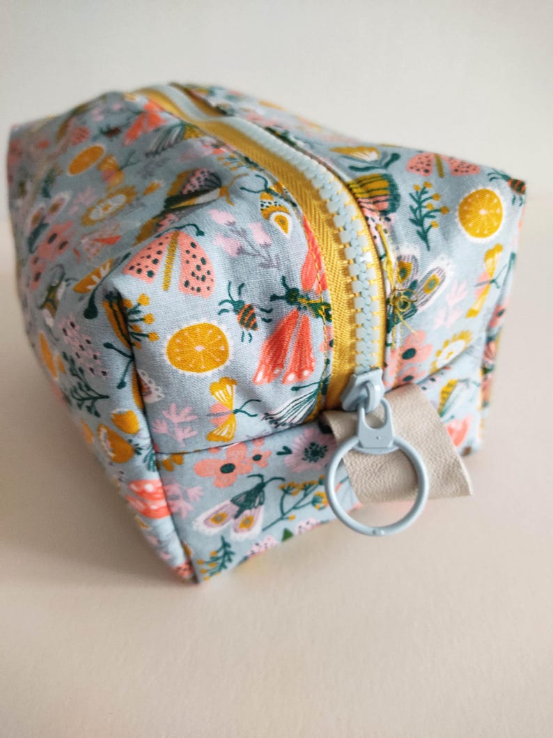 May include: A light blue zippered pouch with a floral and insect print. The pouch has a yellow zipper and a light blue ring attached to a beige strap.