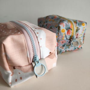 May include: Two small zippered pouches, one with a pink and white abstract pattern and the other with a blue and yellow floral pattern. Both pouches have light blue zipper pulls.