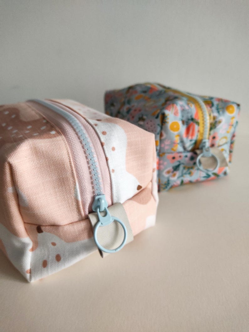 May include: Two small zippered pouches, one with a pink and white abstract pattern and the other with a blue and yellow floral pattern. Both pouches have light blue zipper pulls.