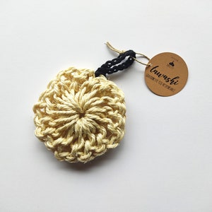 Peut inclure: Un loofah en crochet jaune avec une ficelle noire attachée à une étiquette brune sur laquelle est inscrit "tawashi, CRAFTED FOR GOOD RESPONSIBLY."