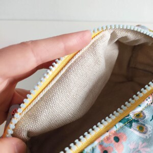 May include: A small, rectangular zippered pouch with a light blue floral print and a yellow zipper. The inside of the pouch is lined with a beige fabric.