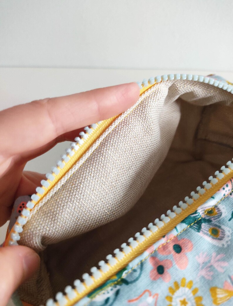 May include: A small, rectangular zippered pouch with a light blue floral print and a yellow zipper. The inside of the pouch is lined with a beige fabric.
