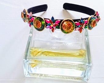 May include: A black headband adorned with colorful gemstones and circular gold-toned accents. The gemstones are in shades of red, pink, green, and blue. The headband is resting on a clear glass block.