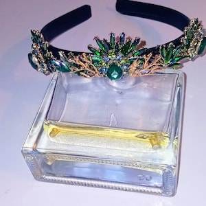 Könnte beinhalten: Ein schwarzes Stirnband mit einem dekorativen Kronenaufsatz aus grünen und blauen Edelsteinen und goldfarbenen Akzenten. Das Stirnband liegt über einem transparenten, rechteckigen Glasbehälter mit einer gelben Flüssigkeit.