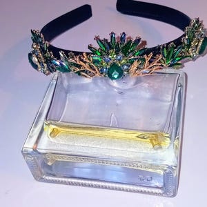 Può includere: Una fascia nera adornata da una corona decorativa con pietre preziose verdi e blu e dettagli color oro. La fascia è posta sopra un contenitore di vetro trasparente e rettangolare con un liquido giallo all'interno.