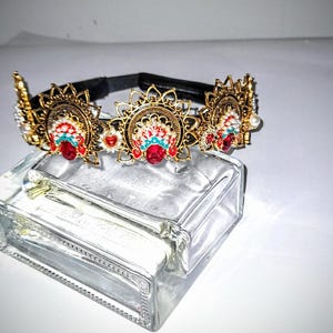 May include: A gold-toned headband with ornate, sunburst-shaped embellishments. Each embellishment features red and turquoise accents, with a central red jewel. The headband rests on a clear glass block.