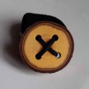 Puede incluir: Un botón de madera con una cuerda negra atada a través de cuatro agujeros en forma de cruz. El botón es de color madera natural y tiene un anillo exterior marrón oscuro.