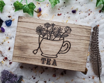 May include: A wooden tea box with a burnt wood finish. The box has a design of a teacup with flowers and the word "TEA" underneath.