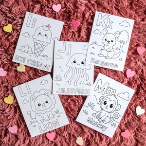 Five white coloring pages with black outlines of cute illustrations. The pages feature the letters I, J, K, L, and M, along with the words "ICE CREAM", "JELLYFISH", "Kangaroo", "LION", and "Monkey". Pink and yellow heart-shaped decorations are scattered around.