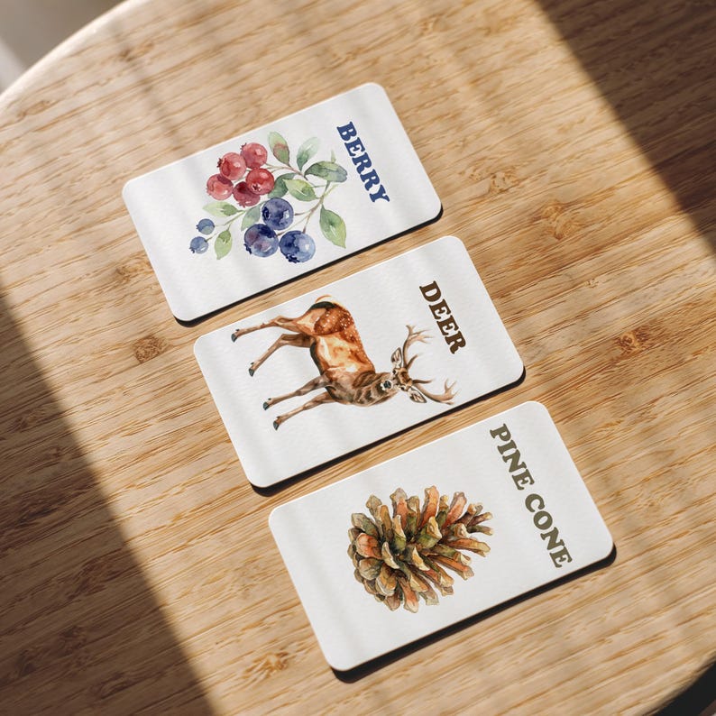 Three rectangular white coasters with nature-themed watercolor designs. One coaster features berries with the word "BERRY", another a deer with "DEER", and the last a pine cone with "PINE CONE".