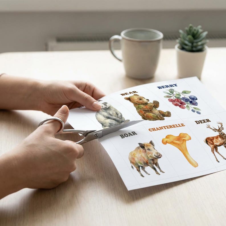 A person is using scissors to cut out illustrated cards featuring watercolor images of a bear, boar, deer, berries, and a chanterelle mushroom. The cards are on a light wood surface.