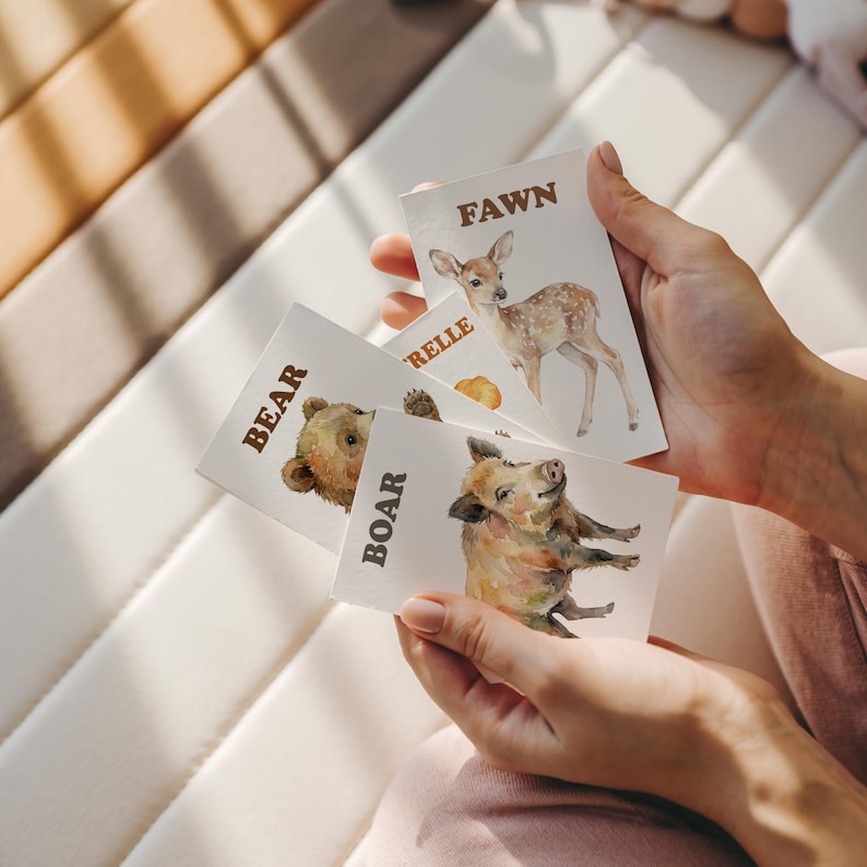 A person holding a set of animal flashcards. Each card features a watercolor illustration of a different animal: a fawn, a bear, and a boar. The animal names are printed in a bold, brown font.