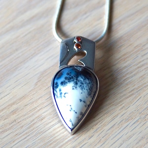 Puede incluir: Un collar de plata con un colgante en forma de l&aacute;grima que presenta una piedra de &aacute;gata blanca y negra. El colgante est&aacute; acentuado con tres peque&ntilde;as piedras naranjas.