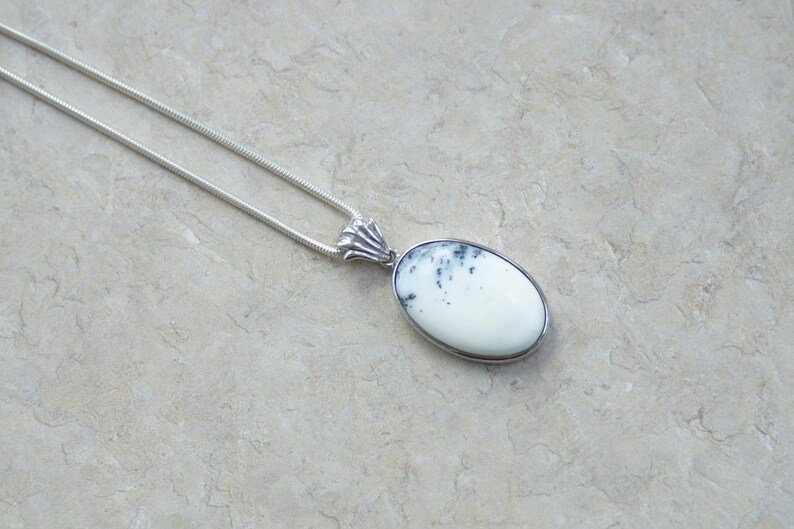 May include: A silver necklace featuring an oval pendant with a white stone and black speckles. The pendant is set in a silver bezel and hangs from a delicate silver chain. The jewelry is displayed on a neutral background.