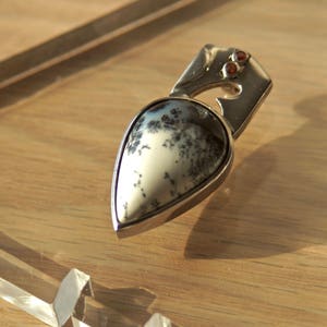 Puede incluir: Un colgante de plata con una piedra de &aacute;gata dendr&iacute;tica en forma de l&aacute;grima engastada en un marco de plata. La piedra tiene un fondo blanco con patrones dendr&iacute;ticos negros. Dos peque&ntilde;as piedras preciosas rojas est&aacute;n engastadas en el marco de plata por encima de la piedra.