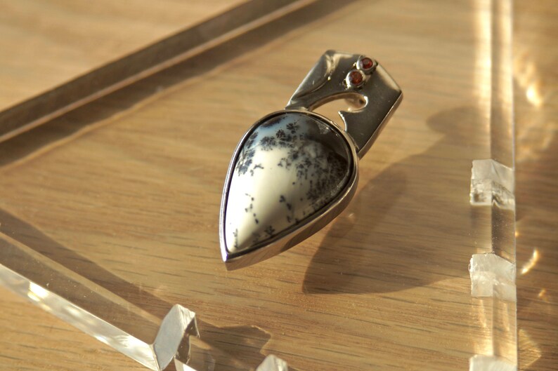 Puede incluir: Un colgante de plata con una piedra de &aacute;gata dendr&iacute;tica en forma de l&aacute;grima engastada en un marco de plata. La piedra tiene un fondo blanco con patrones dendr&iacute;ticos negros. Dos peque&ntilde;as piedras preciosas rojas est&aacute;n engastadas en el marco de plata por encima de la piedra.