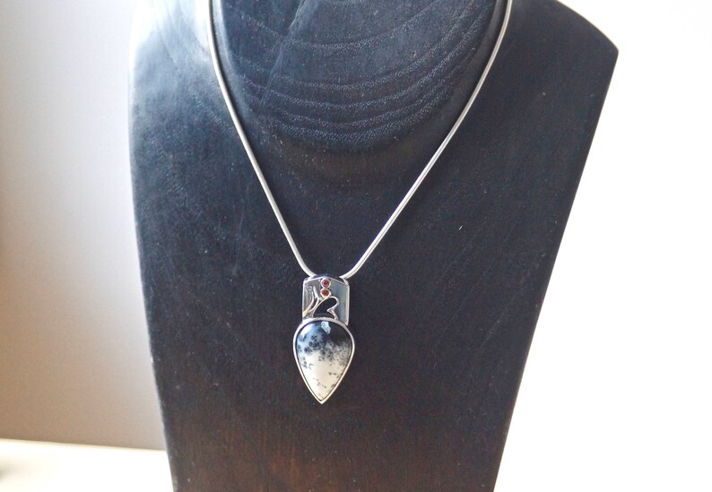 Puede incluir: Un collar de cadena de plata con un colgante en forma de l&aacute;grima con una piedra de &aacute;gata negra y blanca engastada en plata. El colgante tiene un peque&ntilde;o acento de piedra roja.