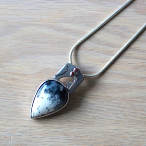 Puede incluir: Un collar de plata con un colgante en forma de l&aacute;grima de &aacute;gata negra y blanca. El colgante tiene un dise&ntilde;o de plata con dos peque&ntilde;as piedras preciosas rojas.
