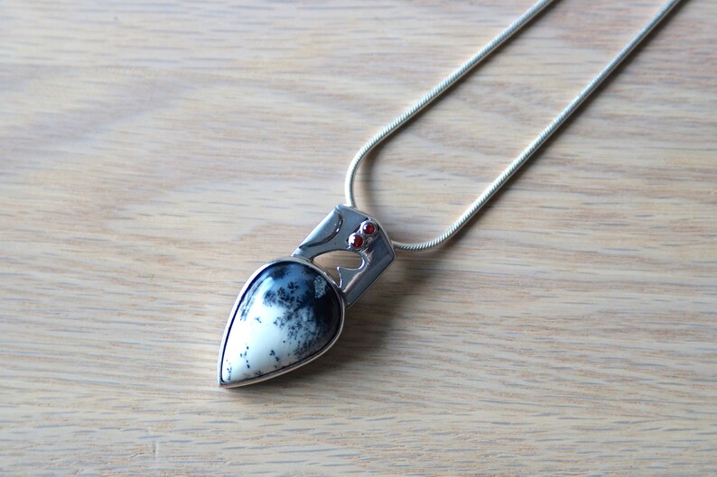 Puede incluir: Un collar de plata con un colgante en forma de l&aacute;grima de &aacute;gata negra y blanca. El colgante tiene un dise&ntilde;o de plata con dos peque&ntilde;as piedras preciosas rojas.