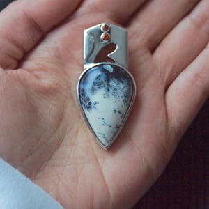 Puede incluir: Un colgante de plata con una piedra de &aacute;gata dendr&iacute;tica en forma de l&aacute;grima engastada en un marco de plata. La piedra tiene un fondo blanco con patrones dendr&iacute;ticos negros. El colgante tiene dos peque&ntilde;as piedras preciosas rojas en la parte superior.