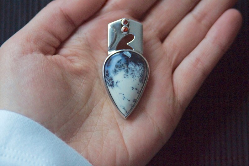 Puede incluir: Un colgante de plata con una piedra de &aacute;gata dendr&iacute;tica en forma de l&aacute;grima engastada en un marco de plata. La piedra tiene un fondo blanco con patrones dendr&iacute;ticos negros. El colgante tiene dos peque&ntilde;as piedras preciosas rojas en la parte superior.