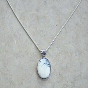 May include: A silver necklace with a white oval pendant featuring black and gray markings. The pendant is set in a silver frame and hangs from a delicate silver chain. The jewelry is displayed on a neutral background.