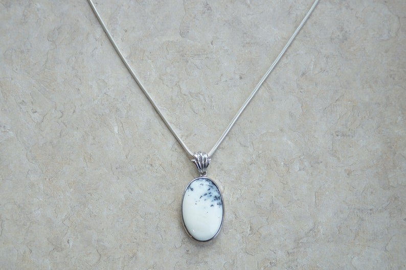 May include: A silver necklace with a white oval pendant featuring black and gray markings. The pendant is set in a silver frame and hangs from a delicate silver chain. The jewelry is displayed on a neutral background.