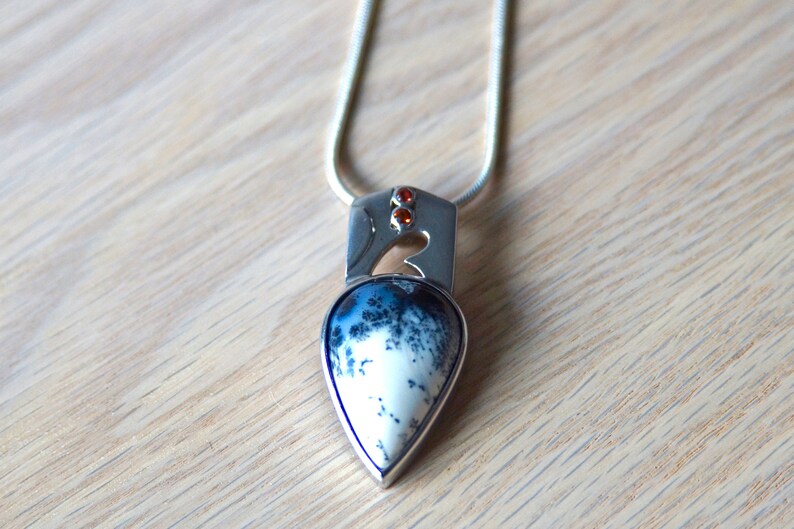 Puede incluir: Un collar de plata con un colgante en forma de l&aacute;grima que presenta una piedra de &aacute;gata blanca y negra. El colgante est&aacute; acentuado con tres peque&ntilde;as piedras naranjas.