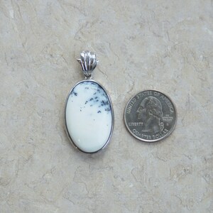 May include: Oval pendant with a white stone and black inclusions, set in a silver-colored metal frame. A quarter coin is next to the pendant for size comparison. The pendant has a decorative bail.