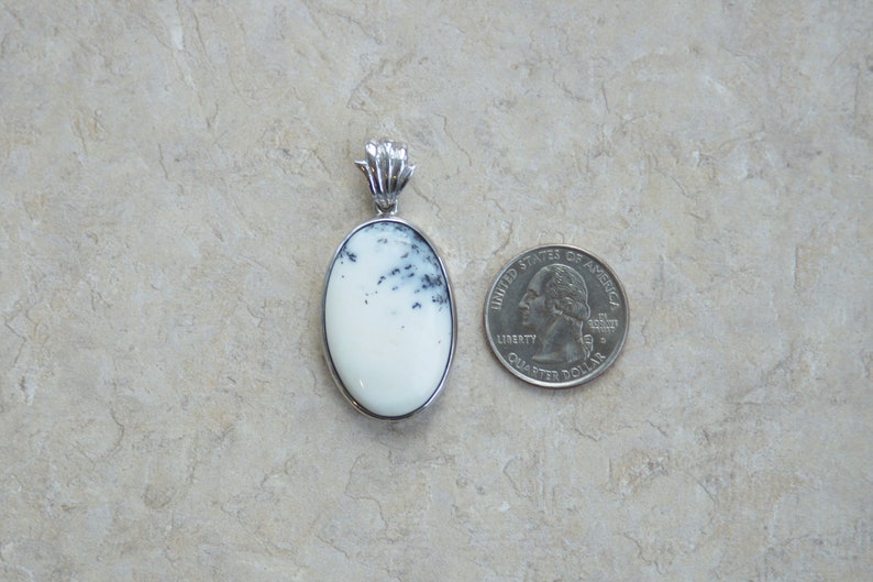 May include: Oval pendant with a white stone and black inclusions, set in a silver-colored metal frame. A quarter coin is next to the pendant for size comparison. The pendant has a decorative bail.