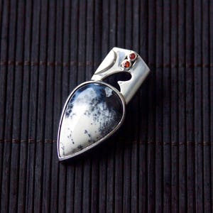 Puede incluir: Un colgante de plata con una piedra de &aacute;gata blanca y negra en forma de l&aacute;grima engastada en un marco de plata. El colgante tiene dos peque&ntilde;as piedras preciosas rojas en la parte superior.