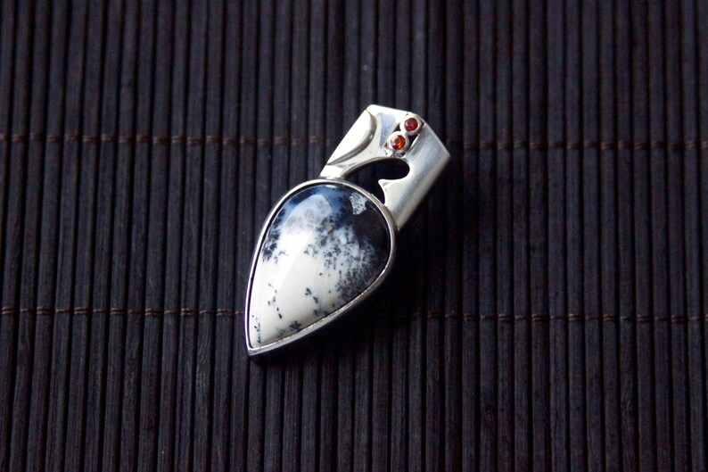 Puede incluir: Un colgante de plata con una piedra de &aacute;gata blanca y negra en forma de l&aacute;grima engastada en un marco de plata. El colgante tiene dos peque&ntilde;as piedras preciosas rojas en la parte superior.