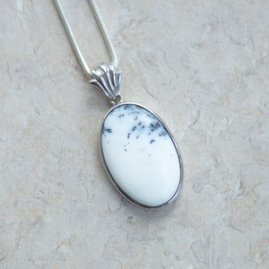 May include: A silver pendant necklace featuring an oval-shaped white stone with black speckles. The stone is set in a silver bezel and hangs from a silver chain. The pendant has a decorative silver bail.