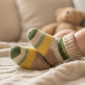 以下が含まれることがあります： 緑、黄色、ベージュのストライプが入ったニットソックスのクローズアップ。靴下は柔らかく、質感のある毛布の上にあります。背景にはテディベアがいます。靴下は手作りのようです。