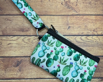 May include: A small, rectangular cactus-themed wristlet with a matching strap. The wristlet features a black zipper and a silver metal ring. The light background is covered with green cacti, pink flowers, and blue accents.