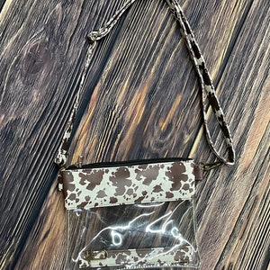 May include: A clear plastic crossbody bag with a brown and white cow print flap. The bag has a long adjustable strap and a zipper closure.