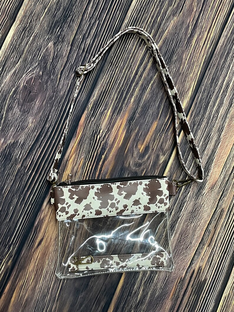 May include: A clear plastic crossbody bag with a brown and white cow print flap. The bag has a long adjustable strap and a zipper closure.