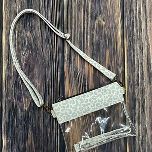 May include: A clear plastic crossbody bag with a white and gray leopard print flap. The bag has a long adjustable strap.