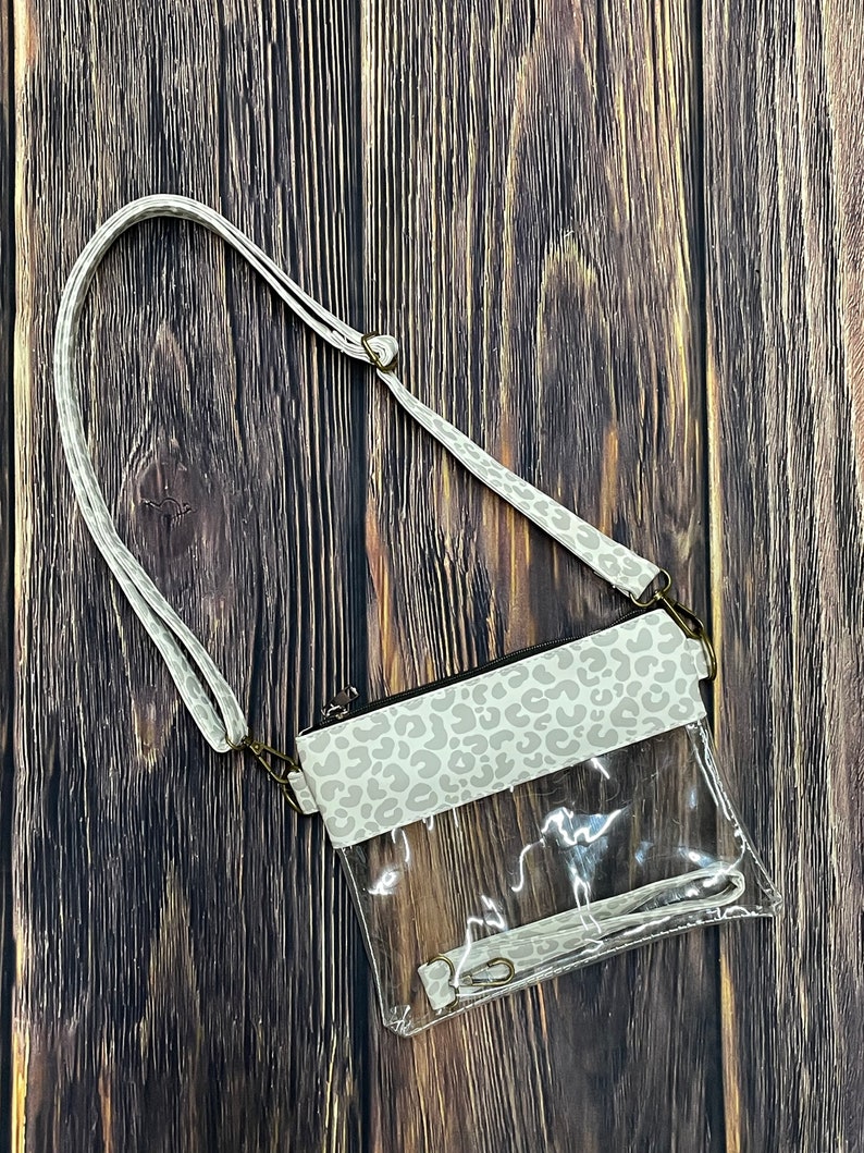 May include: A clear plastic crossbody bag with a white and gray leopard print flap. The bag has a long adjustable strap.