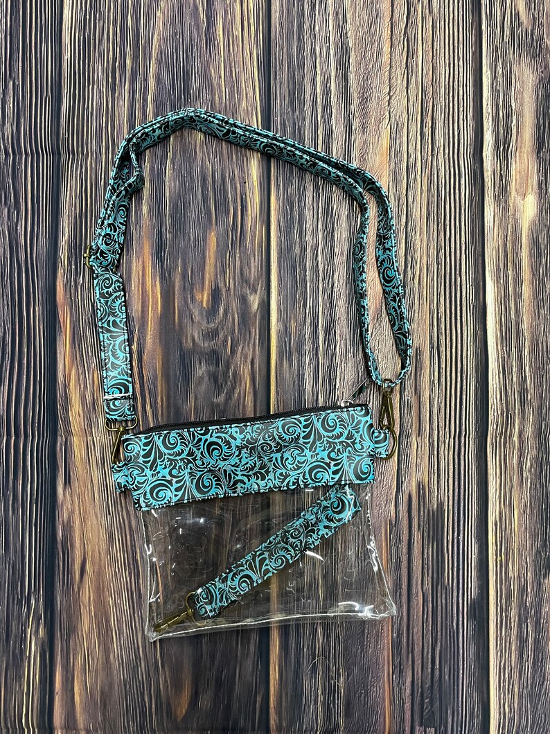May include: Clear plastic crossbody bag with a teal and black patterned strap. The bag has a zipper closure and a smaller clear pouch with a teal and black patterned strap attached to it.