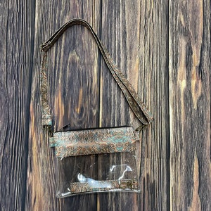 May include: A clear plastic crossbody bag with a brown and teal patterned fabric flap. The bag has a zipper closure and a long adjustable strap.